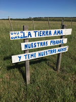 Un cartel pintado de blanco con letras azules, dice: “Utu Guichón: la tierra ama nuestras pisadas y teme nuestras manos”; al fondo hay un alambrado y más atrás una majada de ovejas.