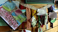 Varias hojas colgadas en una pared de madera, con dibujos en óleo, fotos, un corazón de varios colores y un mensaje escrito, sostenidos por palillos de colgar la ropa.