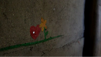 Sobre una pared gris de barro, aparece pintado un pequeño corazón rojo y una flor amarrilla, sobre una línea verde.