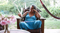 Andrea, una mujer de pelo largo, canoso, con una gran sonrisa, está sentada en un sillón individual de madera en posición de buda vestida con un largo vestido celeste; al fondo se observan árboles y plantas; al frente, una pequeña mesa redonda con una florero y flores rosadas.