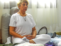 Plano general de Blanca en posición de buda, con sus manos cruzadas y los ojos cerrados, apoyada contra dos almohadones; frente a ella se observan los pies de algunas mujeres que están acostadas.