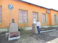 Blanca aparece parada ante un busto de José Pedro Varela que se encuentra junto a otro busto del General Artigas, al frente de una escuela con paredes naranjas.