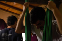 Un hombre joven se sostiene en una tela verde colgada del techo; al fondo se ven otras personas en el interior de una sala con techo de madera.