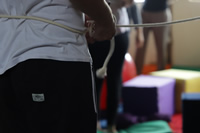 Un hombre joven con pantalón deportivo, se ata una piola blanca a la cintura; al fondo se observan unos almohadones cúbicos de colores y dos mujeres jóvenes de espaldas.