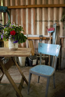 En un galpón de chapas, hay una silla celeste a un costado de una mesa de madera con caballetes sobre la cual hay un jarrón de plástico con flores rojas, blancas y azules.