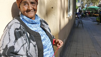 Isabel, una mujer afrodescendiente, bajita y robusta, está parada apoyada contra la pared de una casa; al fondo se observa una feria.