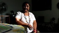 Isabel, con un largo collar de colores, está sentada en una silla en su casa, con un brazo apoyado en una mesa redonda con mantel con flores pintadas.