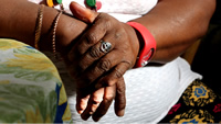 Primer plano de las manos de Isabel, cruzadas una encima de la otra; tiene un par de pulseras de bronce, un reloj rojo y un anillo con el árbol de la vida.