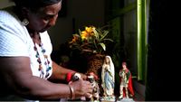 Isabel ordena cuatro pequeñas estatuas en una mesa; una de Santo Expedito, otra de la Virgen, y dos figuras de la cultura afrodescendiente.