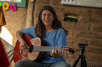 Dentro de una casa de paredes de barro color marrón, Mariana, una mujer de pelo largo color castaño canoso, toca la guitarra y sonríe.