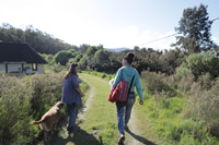 Mariana y Emilia caminan por un sendero que lleva hasta una casa de paredes blancas y techo a dos aguas gris oscuro; a su lado camina un perro de pelo marrón.