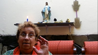 Primer plano de Myriam, hablando y gesticulando con sus manos; al fondo, arriba, en una repisa en la pared, una virgen con un rosario colgado.
