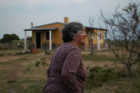 Nelly camina frente a su casa, con su fachada pintada de amarillo; alrededor solo se observa campo.