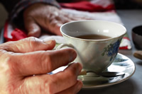Una mano a punto de tomar el asa de una taza con té, pequeña de color blanco con flores pintadas.