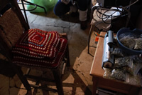 Una silla con un almohadón cuadrado con una funda de lama tejida de colores, frente a una mesa donde hay una máquina para sellar pequeñas bolsas con yuyos.
