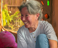 Dentro de una casa de barro, al lado de una ventana por donde ingresa mucha luz, está Olibia, una mujer de pelo plateado recogido, con una gran sonrisa, sentada en el piso con una rodilla sobre su pecho.