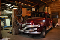 Una vieja camioneta Chevrolet color rojo metálico en muy buen estado, dentro de un galpón donde hay herramientas, tachos de pinturas y otras partes de autos.