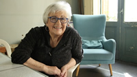 Amparo es una señora con el pelo blanco, corto, con volumen. Usa lentes con marcos azules. Muestra una gran sonrisa. Usa aros plateados y uñas pintadas. Está sentada en una habitación de una casa antigua, se nota por la puerta de fondo con ventanales.