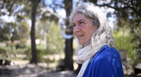 Berta es una mujer adulta. mira a los lejos, tiene su boca entreabierta. Está como distraída con algo que observa. Su pelo blanco ondulado cae sobre su saco azul. Envuelve su cuello con un pañuelo blanco. De fondo hay troncos de árboles altos.