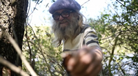 Clever es un hombre mayor con barba larga, espesa y canosa y pelo largo del mismo color. Una boina y unos lentes de marco grueso. Está en medio de unos arbustos; detrás se ve un lago. Extiende su mano y muestra algo a cámara, pero está tan cerca del lente que todo se ve desenfocado. Sonríe.