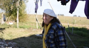Alda usa una boina blanca tejida, camisa de paño a cuadros y una bufanda de lana amarilla. Lleva colgada una cita con llaves. Sonríe y mira a lo lejos. La rodea un paisaje de campo. Está bajo una cuerda con ropa tendida al sol.