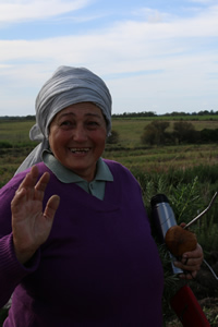 Graciela saluda a cámara con una mano, con la otra sostiene el mate. Bajo el brazo aprieta el termo. Sonríe. Un pañuelo gris, a modo de turbante envuelve su cabeza. De fondo se ve un paisaje de campo típico uruguayo: arbustos bajos y pastizales.