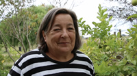 Perla es una señora que sonríe a cámara, con una especie de mueca, y entre cierra los ojos. Tiene el pelo gris, corto y lacio. Detrás de ella un paisaje de campo y un limonero. 
