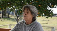 Lila mira a lo lejos, tiene el pelo plateado y corto. La piel arrugada por la edad o por el sol. Detrás hay un naranjo y un paisaje de chacra.