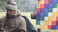 Julio está sentado contra un alambrado, con su mate en la mano y cabizbajo. Usa un gorro de lana tejido. Del mismo alambrado cuelga una bandera de cuadros multicolor, es una Wiphala, bandera de pueblos andinos. 