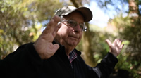 Raúl es un hombre canoso. Usa lentes y un gorro de visera. Está entre árboles con los brazos extendidos y las palmas abiertas, como mostrando el entorno.