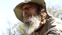 Milton es un hombre adulto. Tiene barba larga y blanca. Pelo largo castaño y entrecanos. Mira a lo lejos. Usa un sombrero de tela. Está en un lugar arbolado y con cielo despejado.