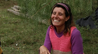 Claudia es una mujer adulta pero no mayor. Tiene el pelo castaño sostenido por una vincha de colores. Lleva puesto un chaleco tejido de colores vivos. Sonríe y cierra sus ojos. Está sentada bajo un sauce.