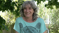 Sylvia sonríe. Su pelo gris y ondulado cae sobre sus hombros. Viste un buzo verde con diseños de mandalas. Está a la sombra de un árbol y rodeada de vegetación.